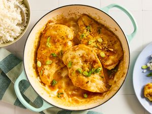 Overhead view of a teal dutch oven with three honey soy chicken breasts and sauce topped with green onions next to a bowl of white rice, a striped fabric napkin and blue plate of recipe and chicken