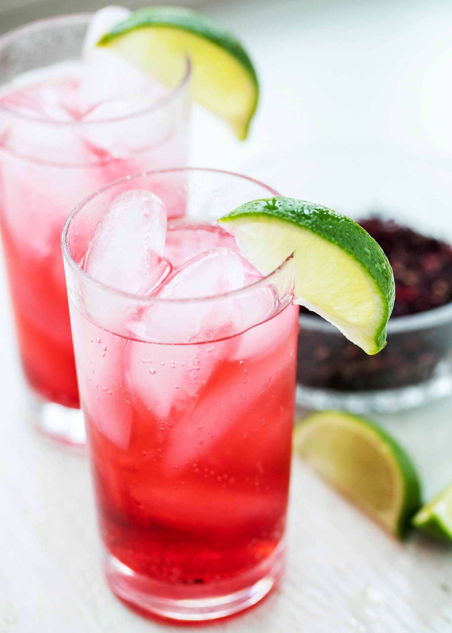 Two tall glassed with hibiscus mocktails inside. Ice cubes are inside the glass and a lime wedge sits on the lip of each glass with additional lime wedges next to the glass in front. Partial view of a bowl of dried hibiscus in the background to the right of the second glass.