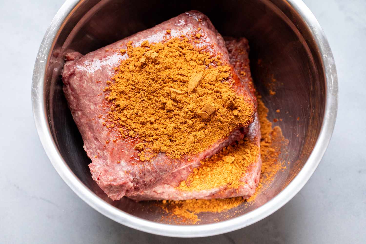 Ground beef topped with seasonings in a metal bow.