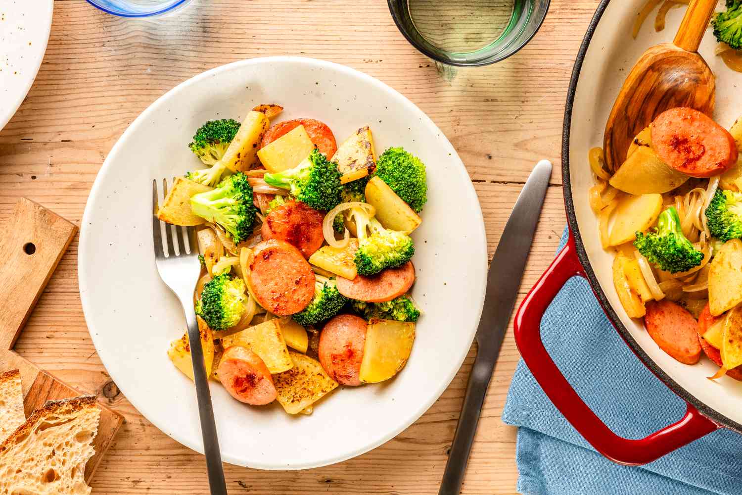 Overhead view of a white bowl of Kielbasa, Potato, and Broccoli Skillet with a fork next to a knif,e a skillet, bread on a cutting board and a drinking glass 