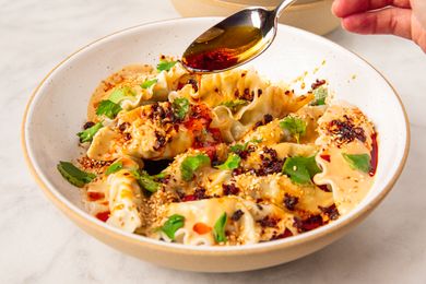 Dumplings on a plate topped with peanut chili sauce and garnished with sesame seeds and herbs with a hand pouring chili oil from a spoon