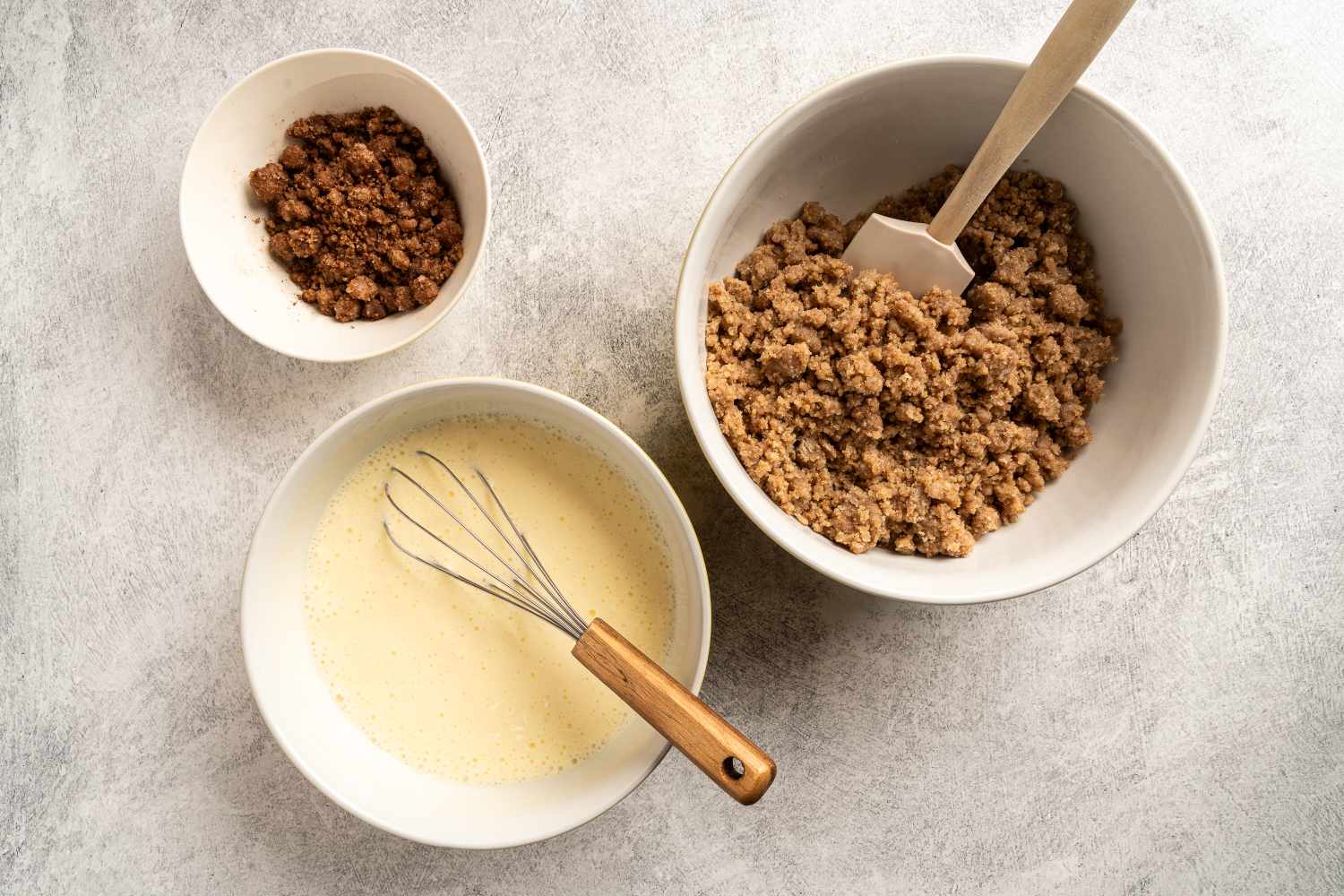 Three bowls on a countertop containing ingredients for a recipe a whisk and a spatula are shown