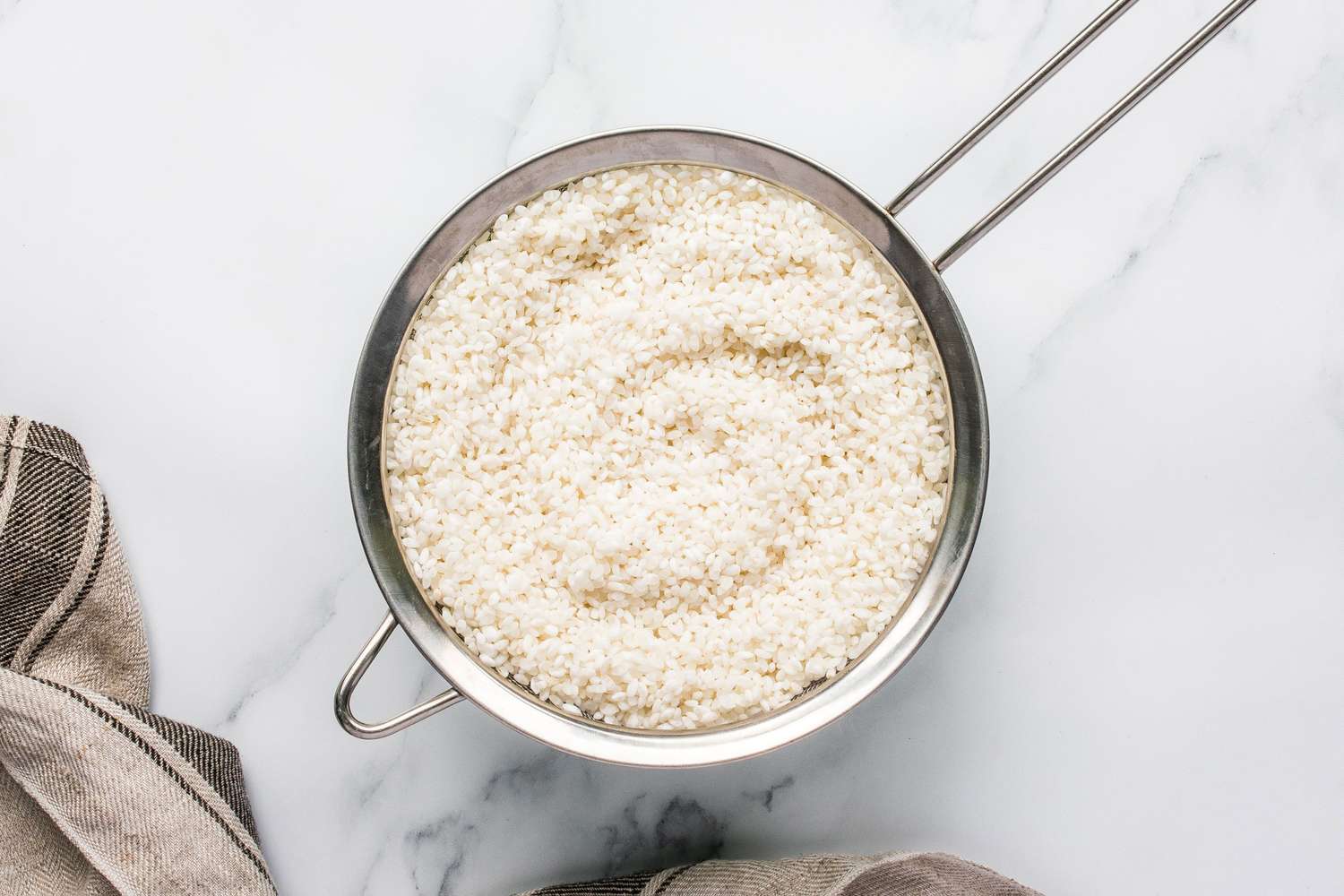 A bowl of rinsed rice sitting in a fine mesh strainer placed on a light surface with a towel nearby