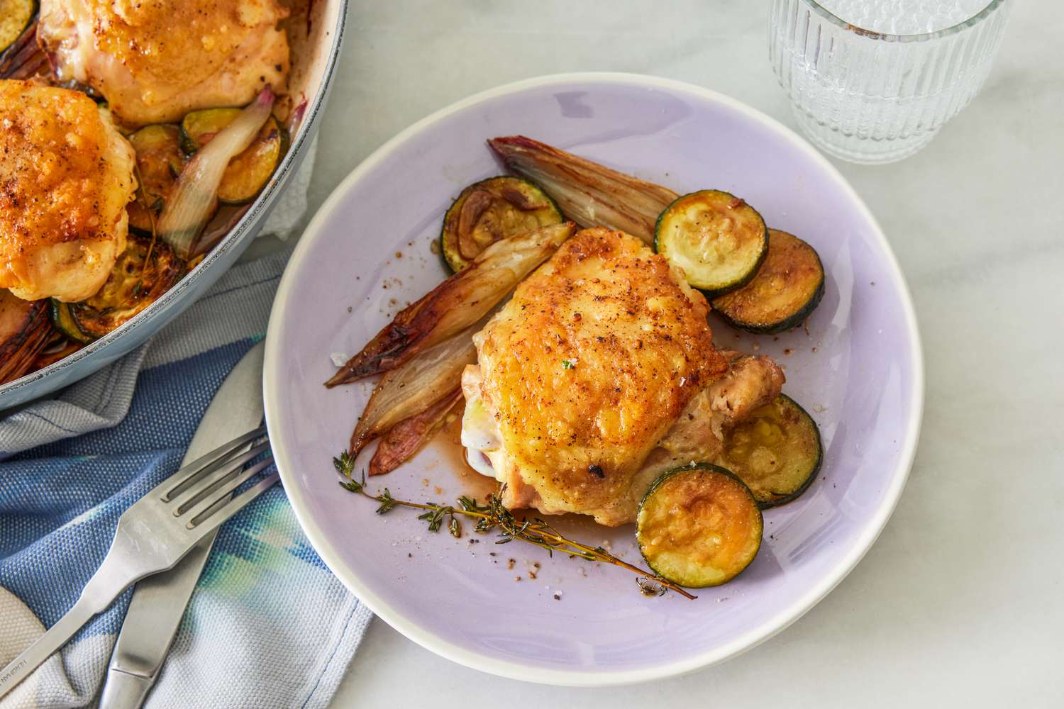 serving of crispy chicken thighs with zucchini and shallots on a plate at a table serving with a dutch oven (with more chicken), a glass of water, and utensils on a kitchen napkin