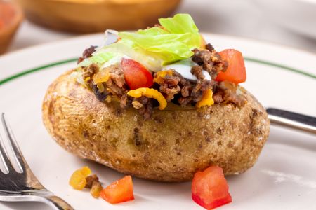 A baked potato topped with ground meat cheese tomatoes and lettuce on a plate with a fork nearby
