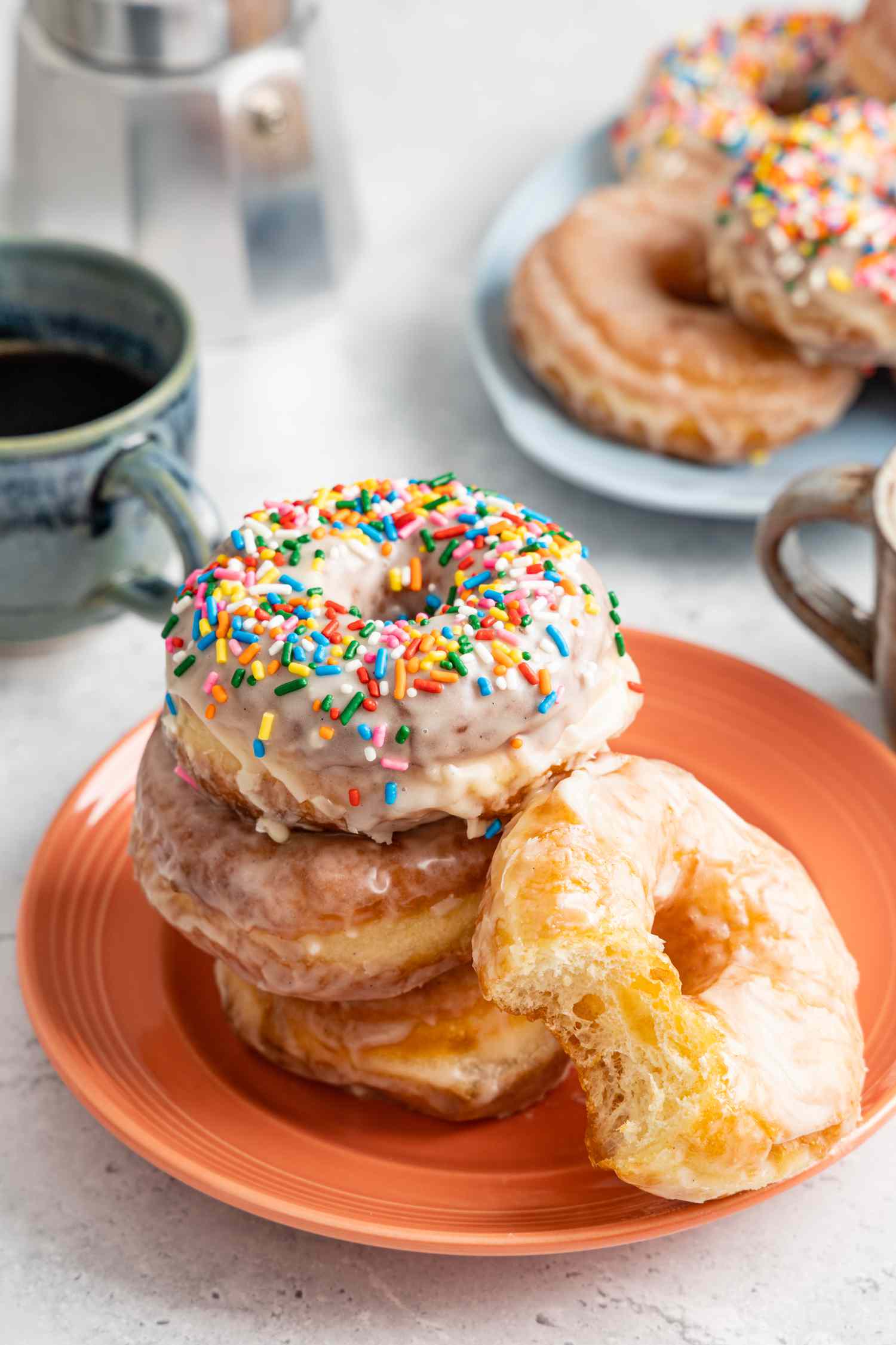 Stack of Glazed Donuts with the Top Donut Covered in Sprinkles, and Leaning Next to the Stack, a Glazed Donut with a Bite Missing, All on a Platter. In the Background, a Two Mugs of Coffee and Another Platter of Glazed Donuts