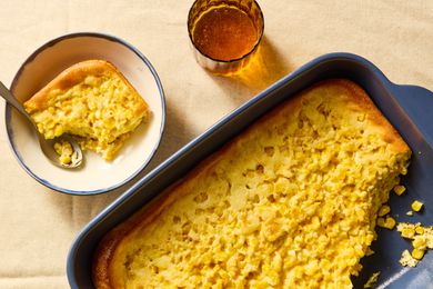 Overhead view of a dark blue baking dish of corn soufflé with a serving removed and a white bowl and spoon with a serving next to a drinking glass