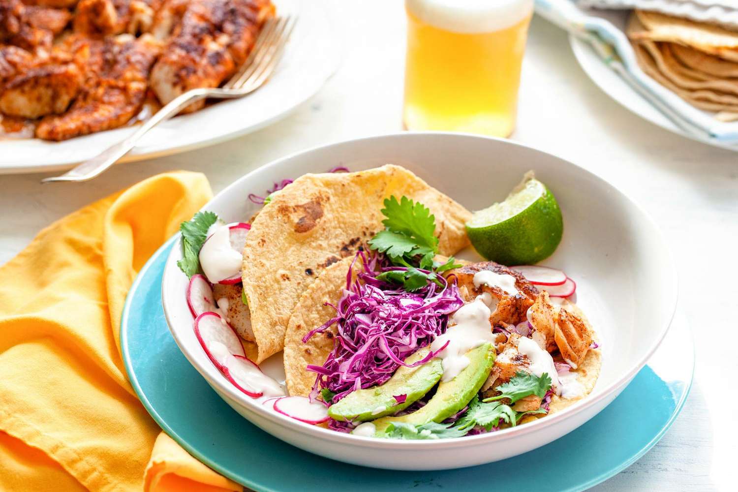 A table is set with a plate with fish tacos. Corn tortillas layered with fish, avocado, cabbage, cilantro, cheese and lime.