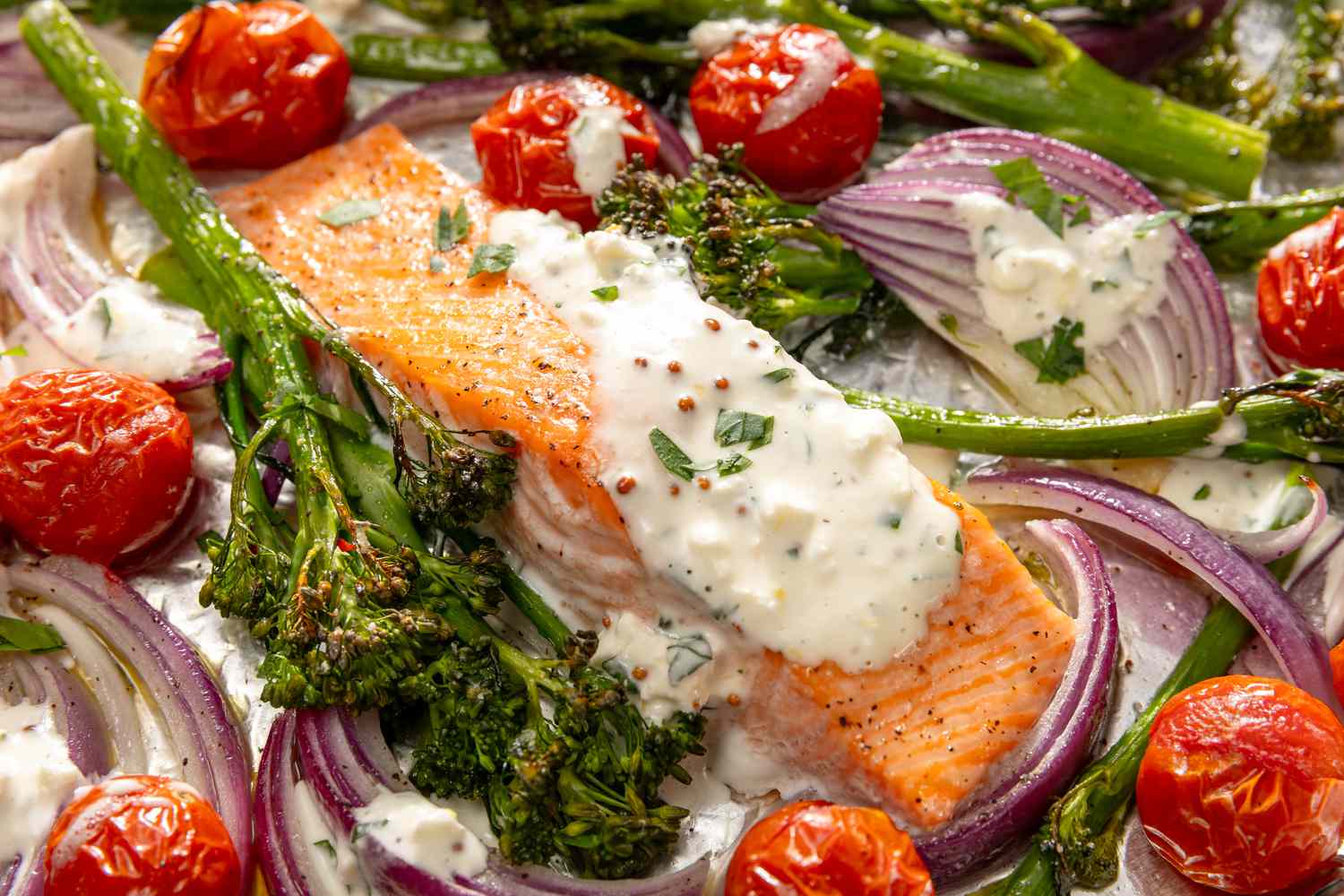 Close up view of a piece of salmon with feta sauce along with broccolini, cherry tomatoes and onions