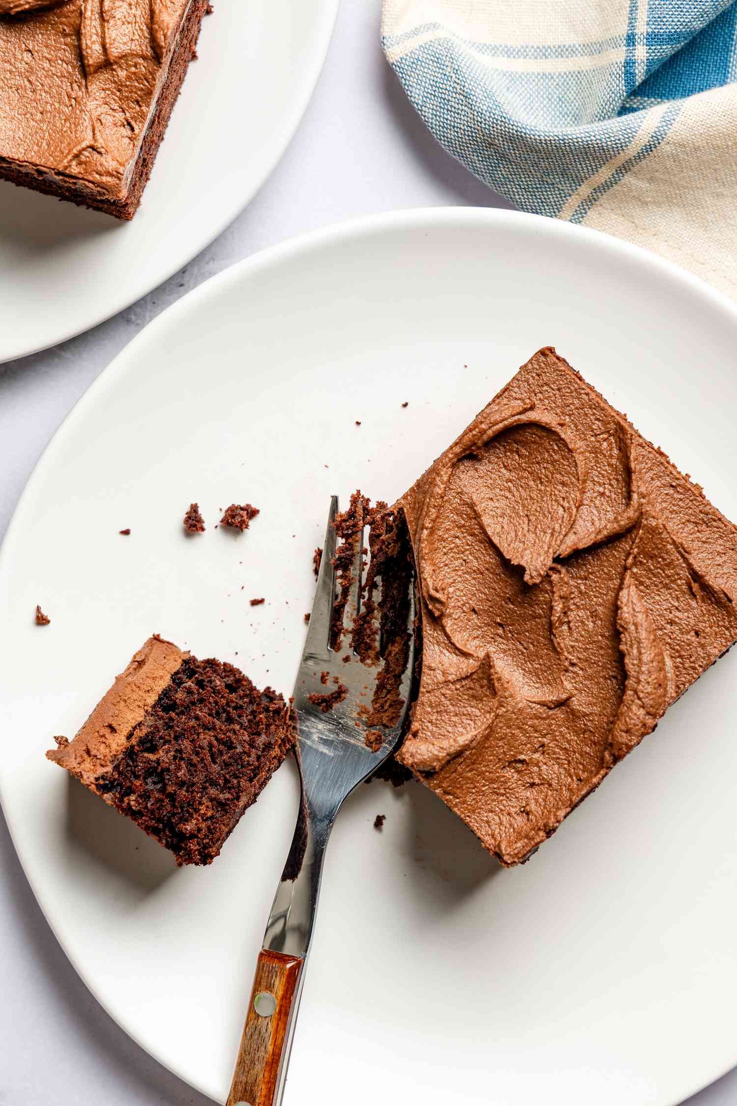 slice of devil's food cake on a plate with a piece broken off using a fork, and in the surroundings, another slice of cake on a plate and a table napkin