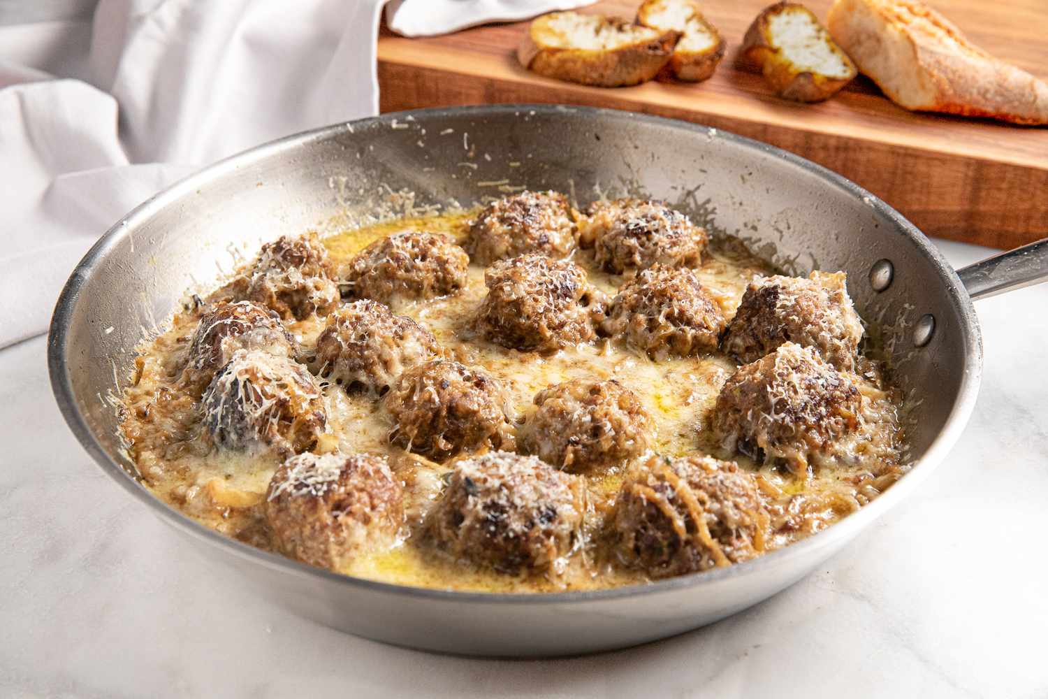 French Onion Meatballs in a Pan, and in the Background, Toasted Baguette Slices on a Wooden Platter and a White Table Cloth Draped Onto It