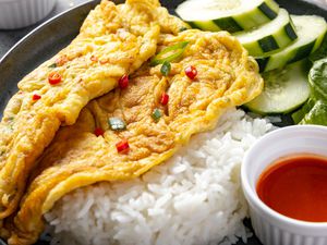 Close Up: Plate of Kai Jeow (Thai Omelet) with Cucumber Slices, Romaine Leaves, Rice, and a Small Bowl of Sriracha