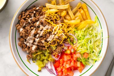 A deconstructed burger bowl with ground beef shredded cheese lettuce tomato onions and fries