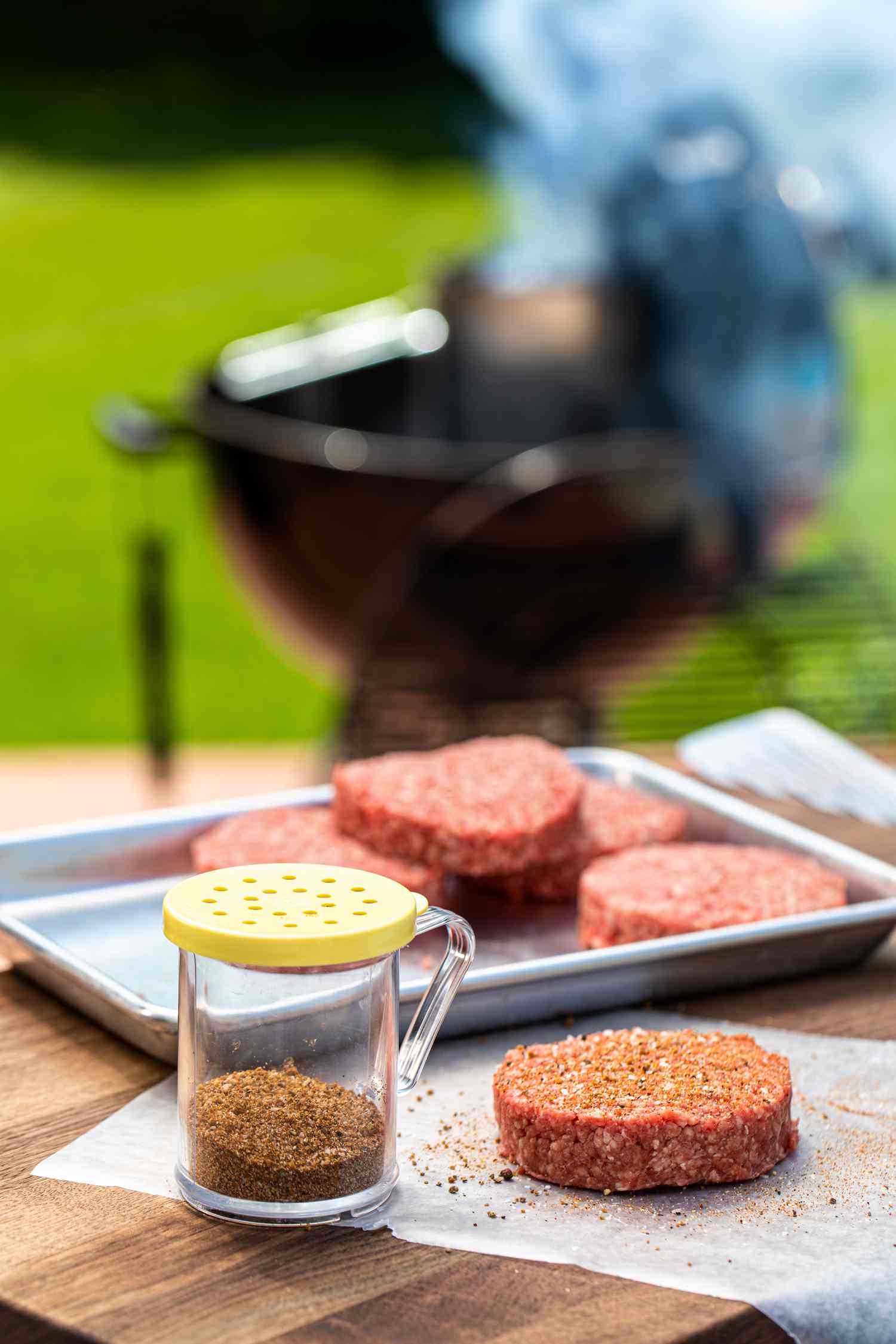 Hamburger patties set next to a jar of the best hamburger seasoning and a grill in the background.