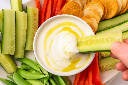 A bowl of feta dip surrounded by sliced vegetables and crackers with a hand dipping a cucumber stick into it