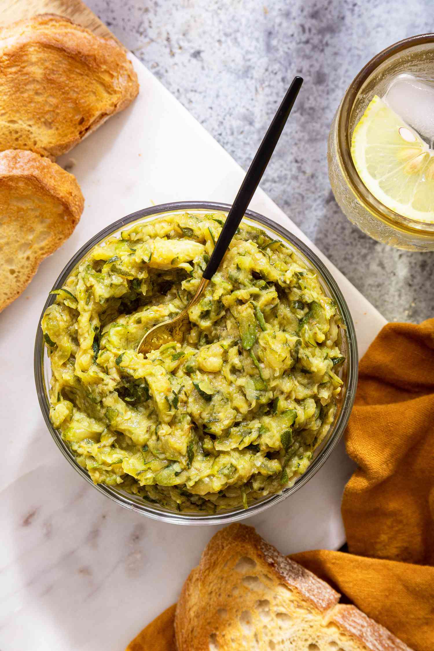 Bowl of Zucchini Butter With a Spoon on a Marble Slab With Toast, and in the Surroundings, a Mustard Colored Kitchen Towel and a Glass of Water