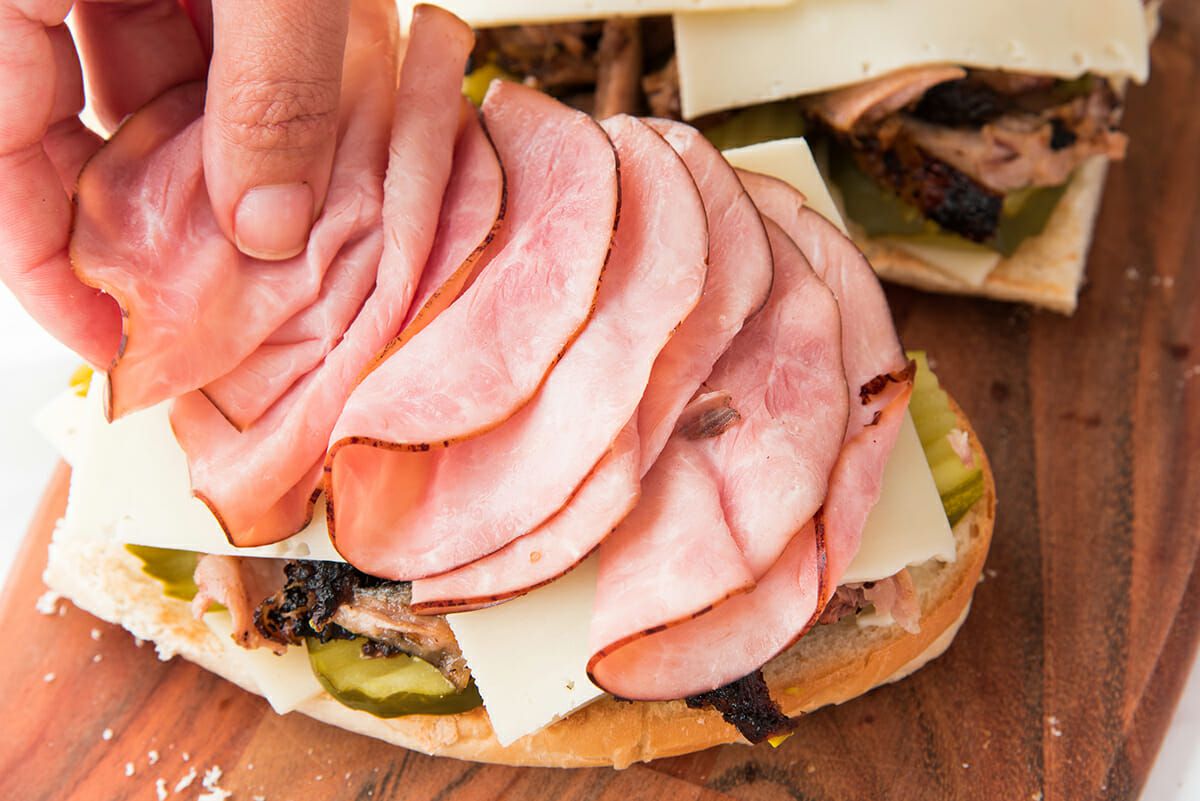 Sliced french bread set on a cutting board with a hand placing sliced ham on the sandwich, along with pickles, cheese and pork. Second half of the sandwich is visible in the right corner.
