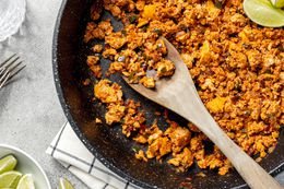 Copycat Sofritas in a Pan with a Wooden Spoon and Lime Wedges, Surrounded by a Bowl of Lime Wedges, a Glass of Water, and a Couple of Forks