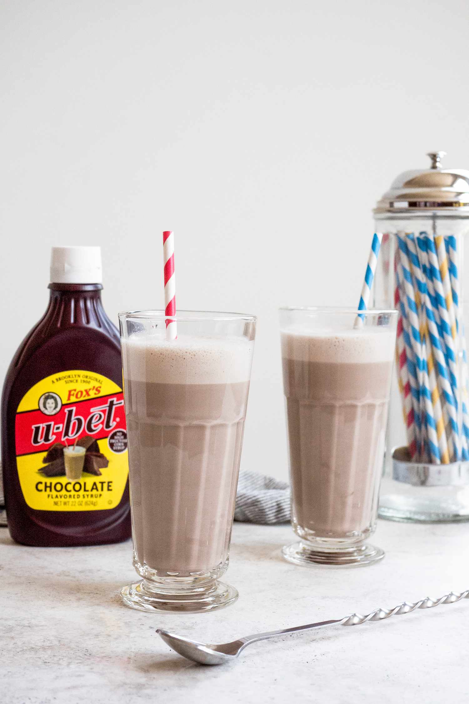 Classic Egg Cream in Two Glasses with Bottle of Chocolate Sauce and Container of Straws in Background