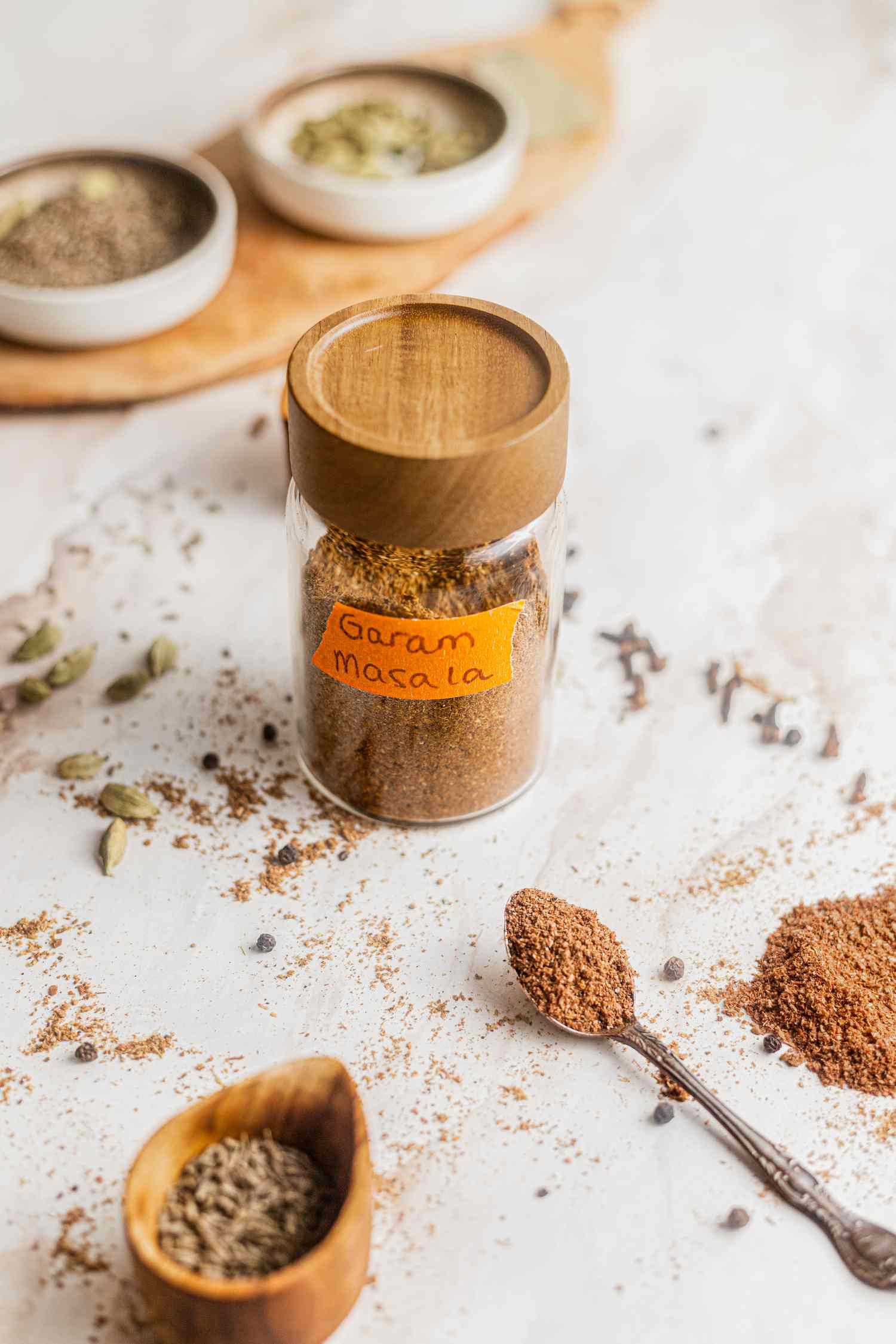 Labeled Jar of Garam Masala Surrounded by Whole Spices and Garam Masala on the Counter, a Spoonful of Garam Masala, a Pile of Garam Masala, and Bowls With Whole Spices 
