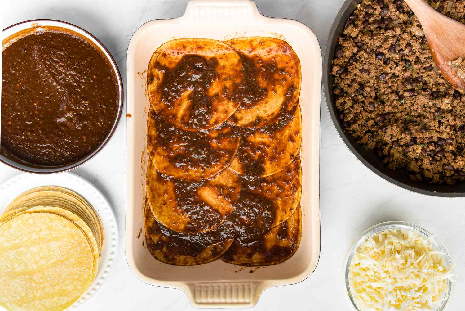 Casserole Dish with Enchilada Sauce Soaked Tortillas Surrounded Enchilada Ingredients (a Plate of Tortillas, Bowl of Sauce, Bowl of Shredded Chicken, and Bowl of Shredded Cheese)