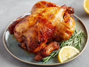 A crispy skinned roasted chicken on a plate, with rosemary sprigs and a lemon wedge as garnish