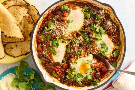 A round baking dish with the black bean and egg bake recipe, next to a plate of blistered corn tortillas and garnish of cilantro and fresh cheese