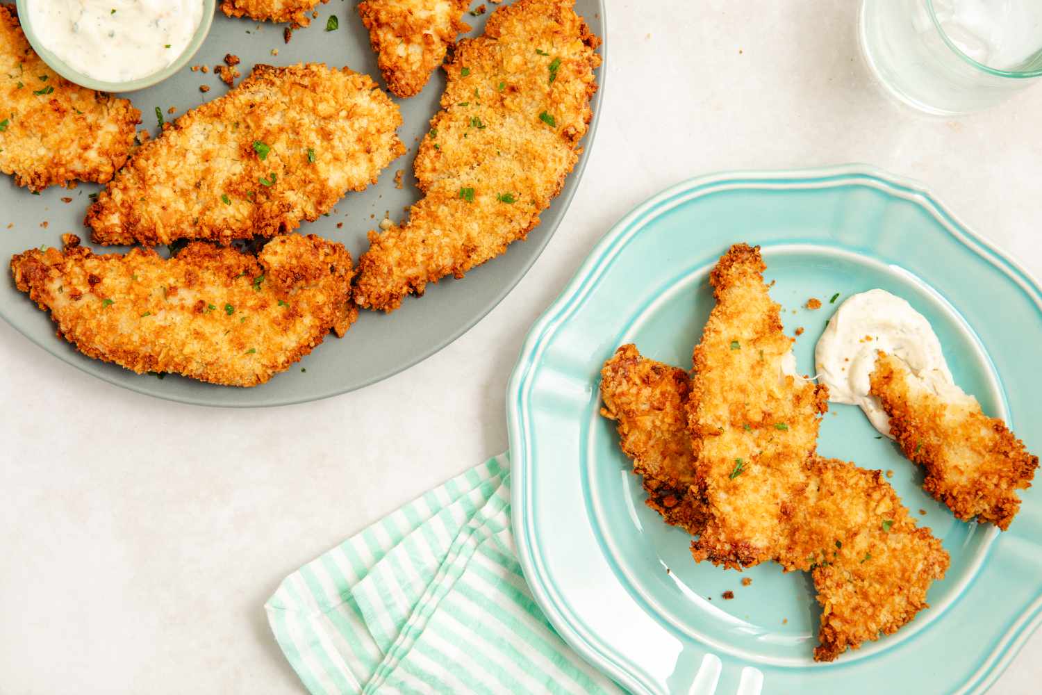 Air-fryer chicken tenders on a plate with some ranch next to a platter with more chicken tenders and a bowl of ranch, all at a table setting with some kitchen towels and a glass of water