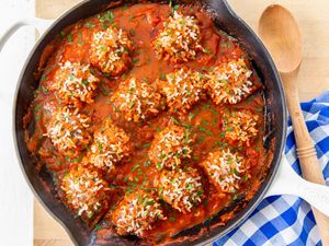 Overhead view of a cast iron skillet of meatballs with expanded rice on the surface and sauce on a wooden cutting board next to a checkered blue and white towel and wooden spoon