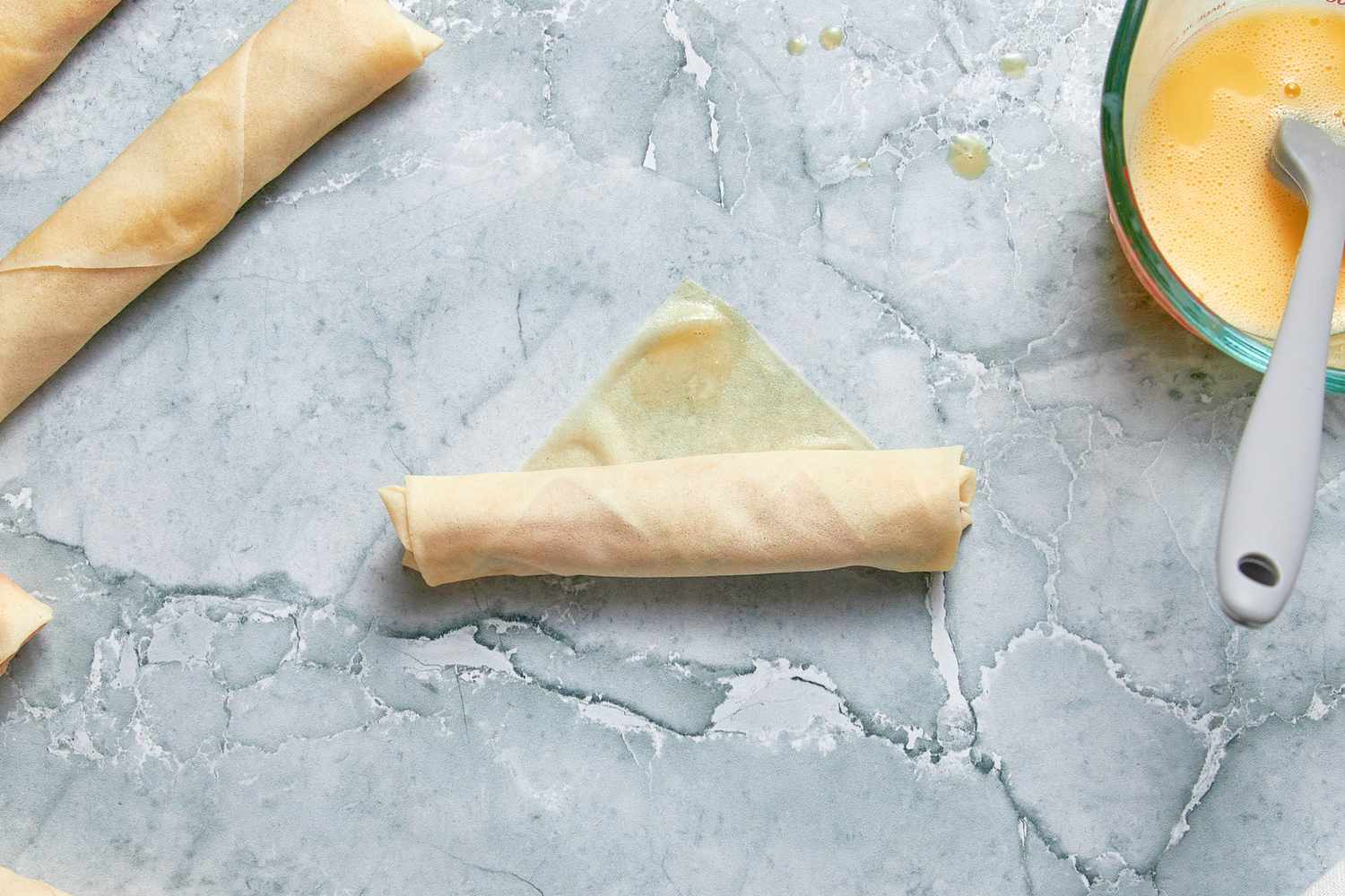 Lumpia Rolled Upwards Towards the Top Corner of the Egg Roll Wrapper, and in the Surroundings, and in the Surroundings, Rolled Lumpia and a Pyrex Measuring Cup With the Egg Wash