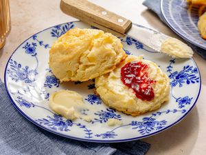 Angled view of a decorative small plate with one sliced mashed potato biscuit with butter and jam next to a butter knife with butter on the end