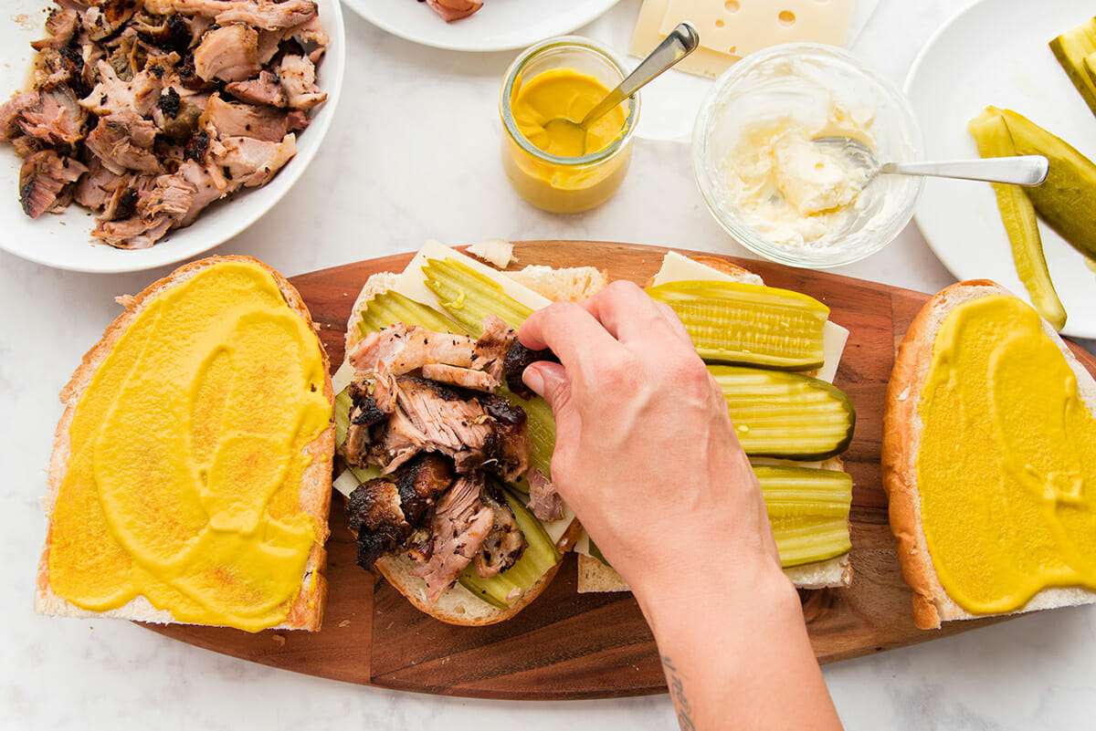 Best cuban sandwich is being assembled on a wooden cutting board set on a marble countertop. Hand is placing shredded pork, along with picles and cheese on french bread. Two tops of the bread are covered with yellow mustard and bowls of pork, mustard, mayonnaise and pickles are visible.