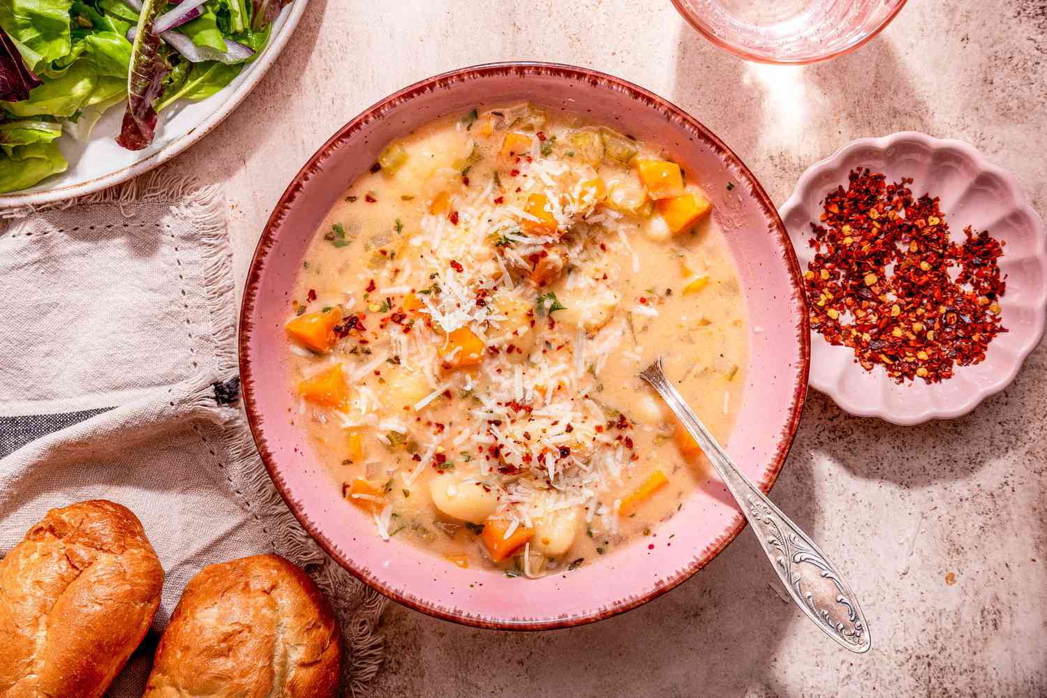 Bowl of creamy vegetable dumpling soup with a spoon, and in the surroundings, a plate of salad, a glass of water, a small bowl of rushed peppers, and dinner rolls on a grey and black kitchen towel