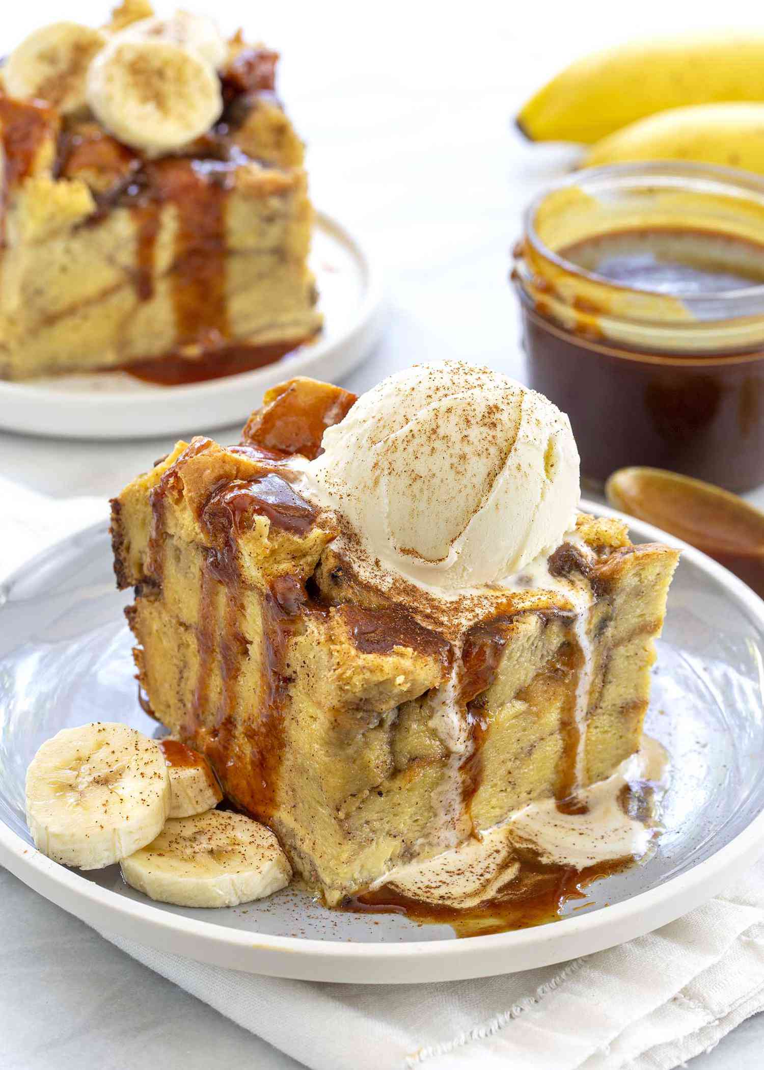 Slice of the best slow cooker banana bread pudding topped with a scoop of ice cream and drizzeld with caramel sauce on a gray plate. A jar of caramel sauce is off to the side.