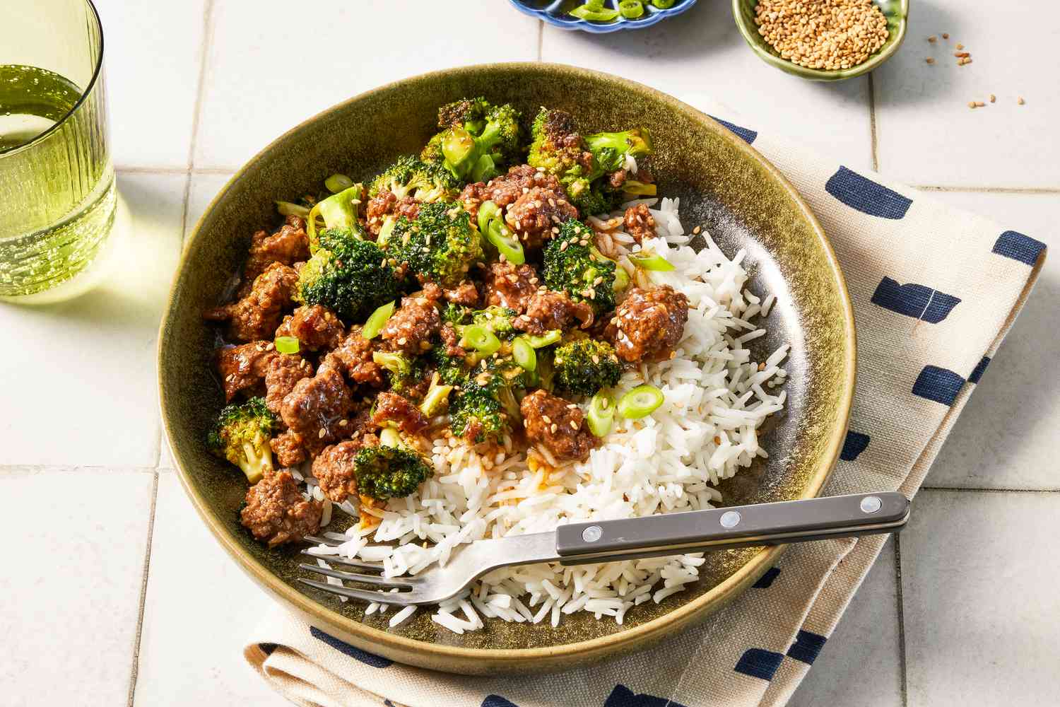 Bowl of ground beef and broccoli stir fry with a serving of rice at a table setting with a bowl of sesame seeds and a glass of water