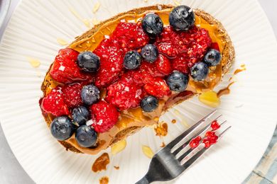 Slice of toast topped with peanut butter, raspberries, and blueberries, placed on a white plate with a fork to the side