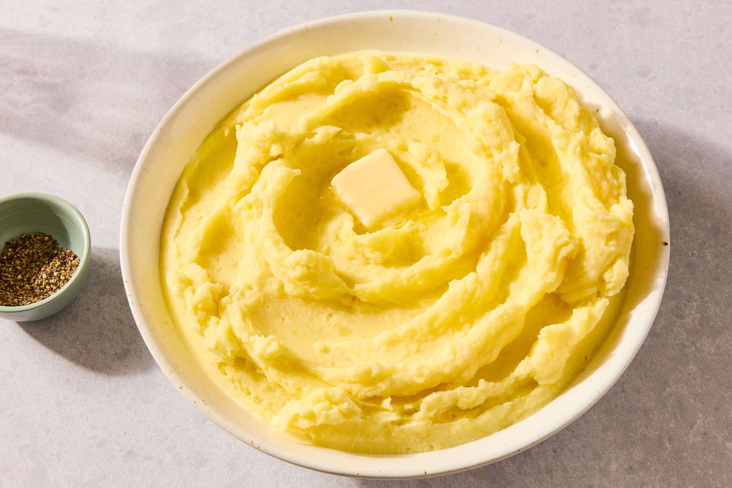 large bowl of mashed potatoes with a pad of butter on top, small bowl with black pepper