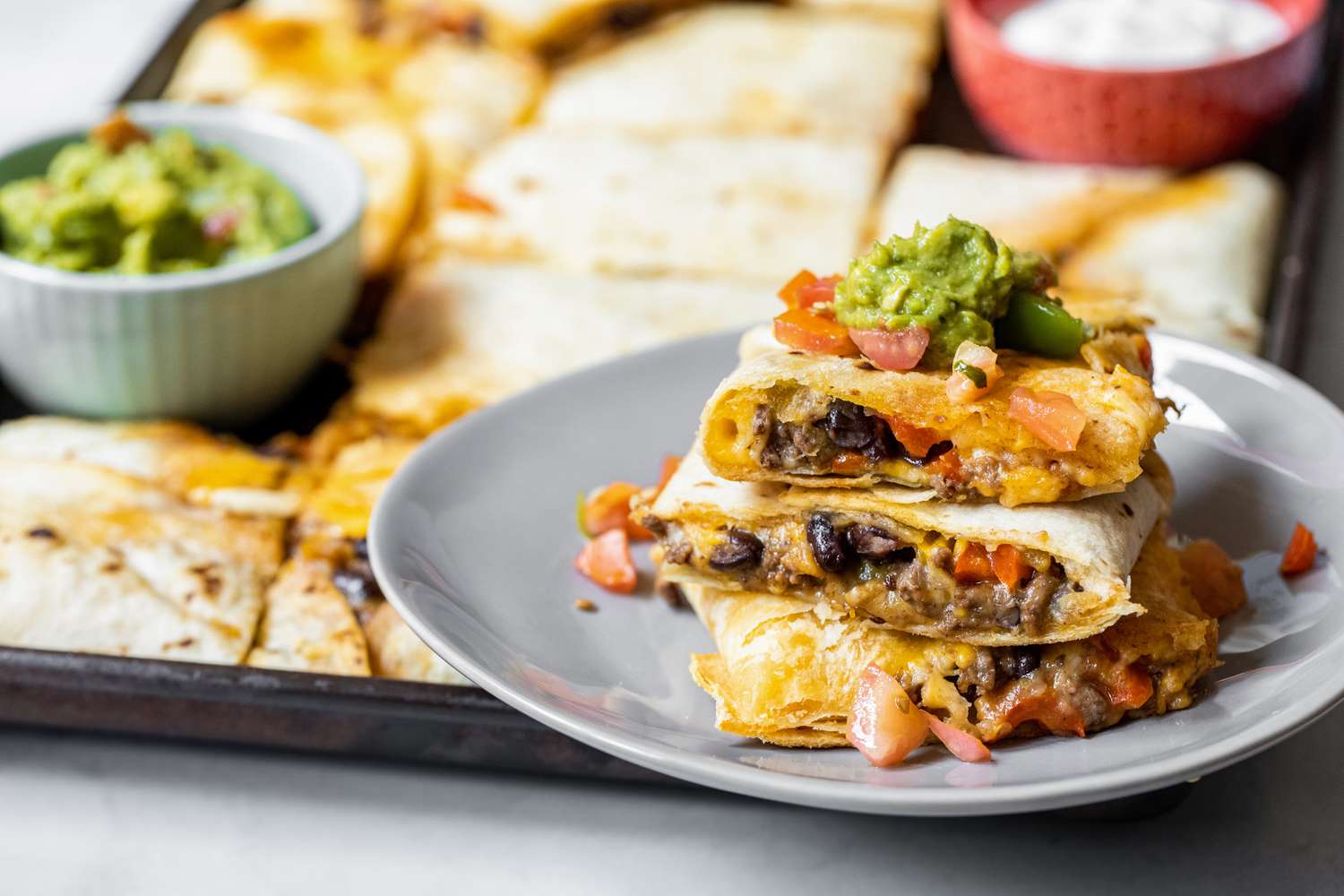 Stack of Loaded Sheet Pan Quesadillas Pieces on a Plate Topped with Salsa and Guacamole. More Quesadilla on the Sheet Pan in the Background.