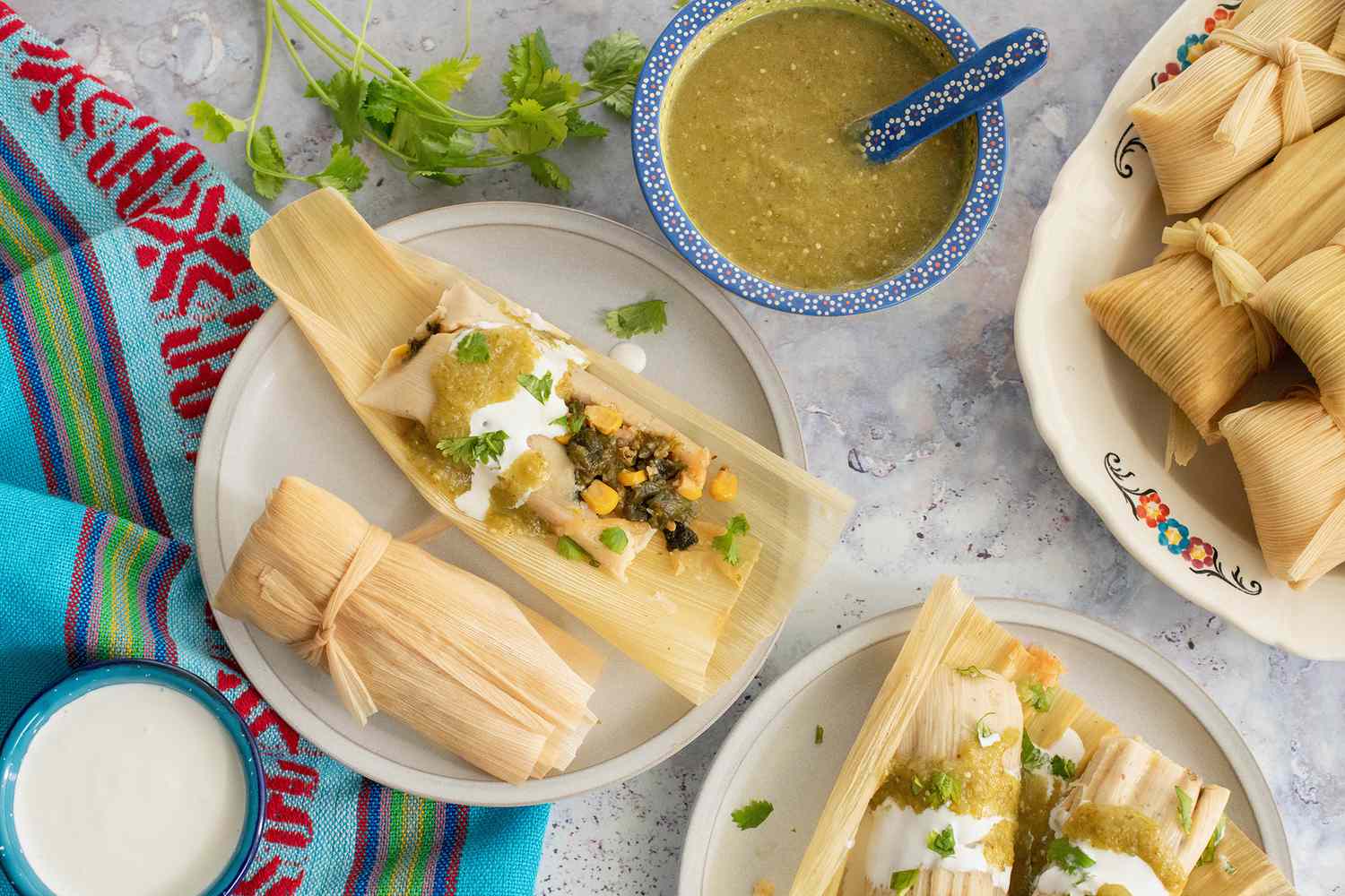 Poblano, Corn, and Cheese Tamales Topped with Salsa Verde, Crema, and Cilantro on Two Plates and Surrounded by a Bowl of Salsa Verde, a Bowl of Crema, and a Platter with More Tamales All on a Table Runner