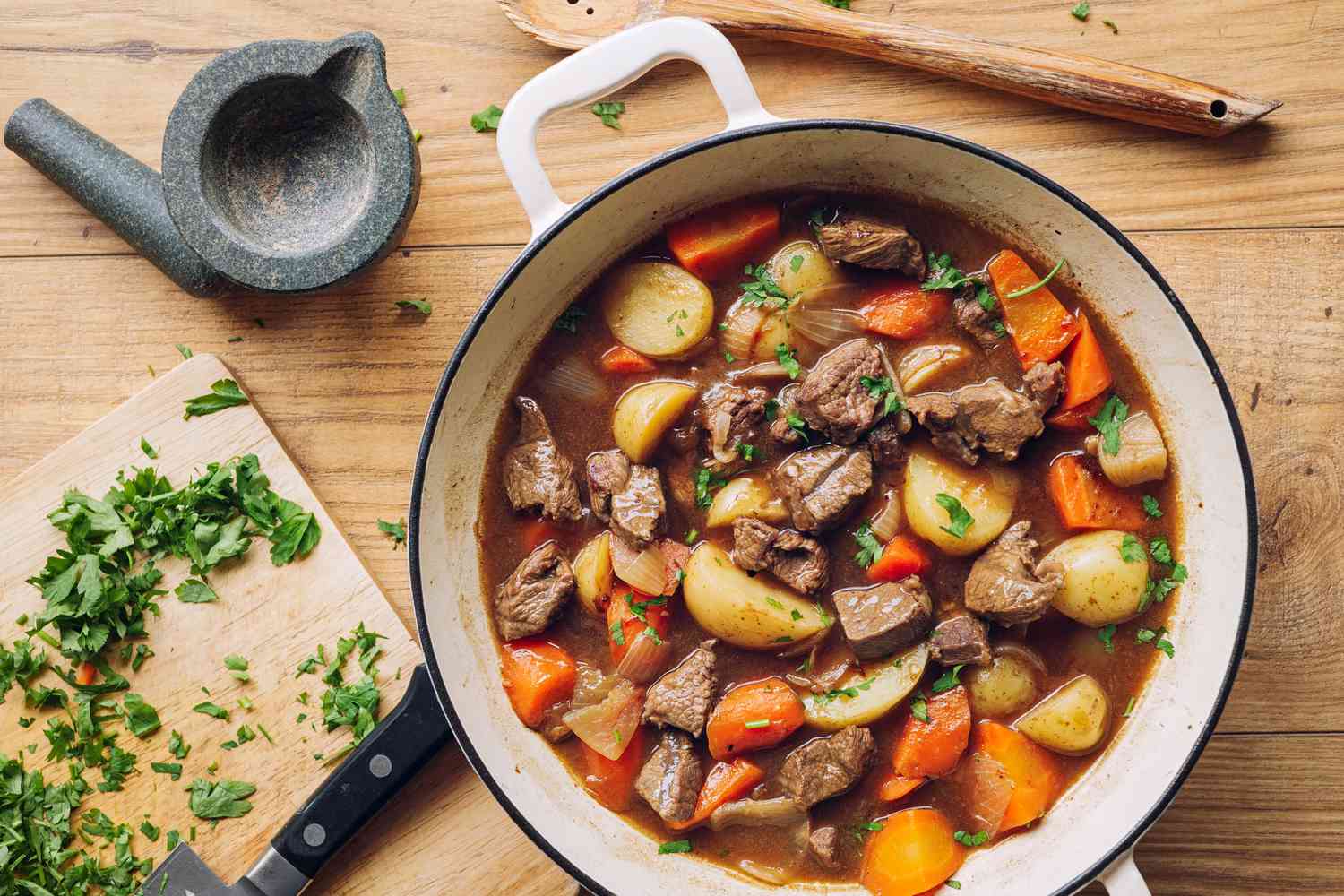 Overhead shot of a Dutch oven with beef stew on a wooden surface, with chopped parsley on a cutting board on the side
