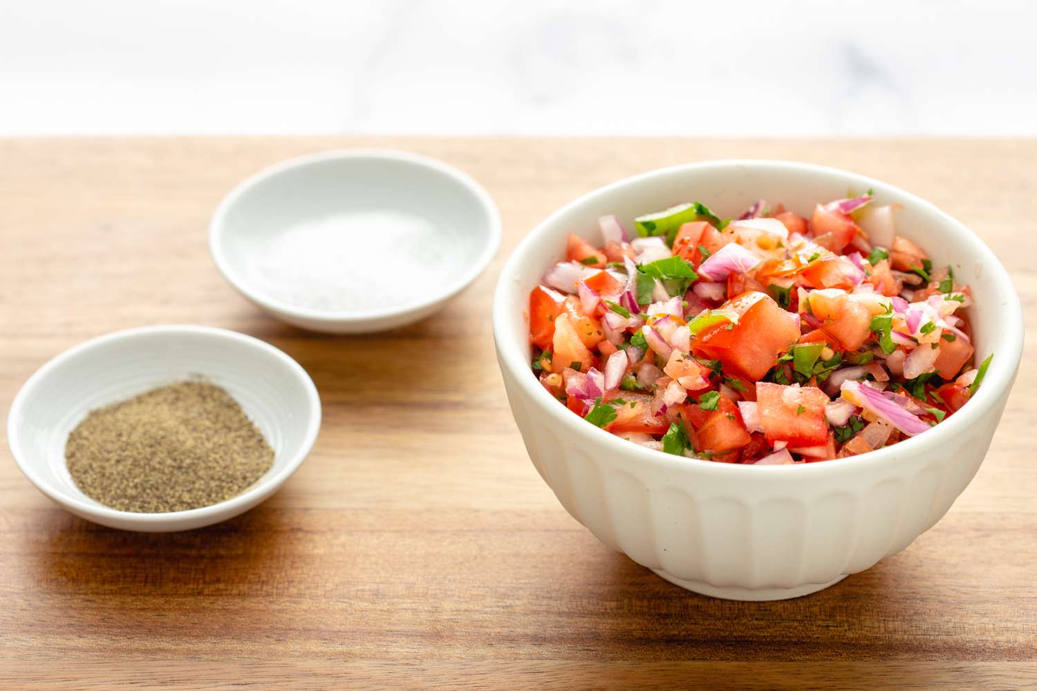 A bowl with salsa in a white bowl with salt and pepper next to it.