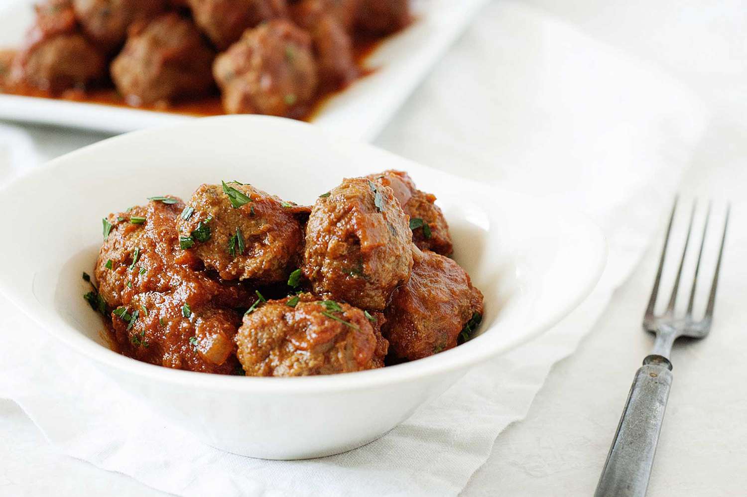 A bowl of meatballs with a platter of meatballs behind it.