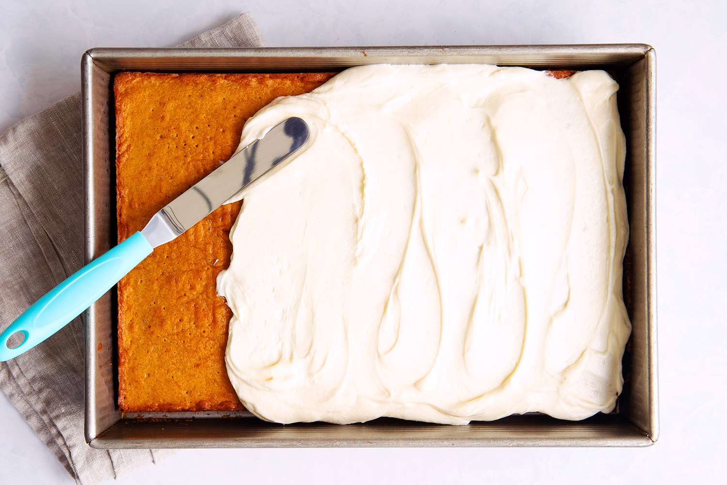 A rectangular cake being frosted with a cream spread in a pan a bluehandled knife is used