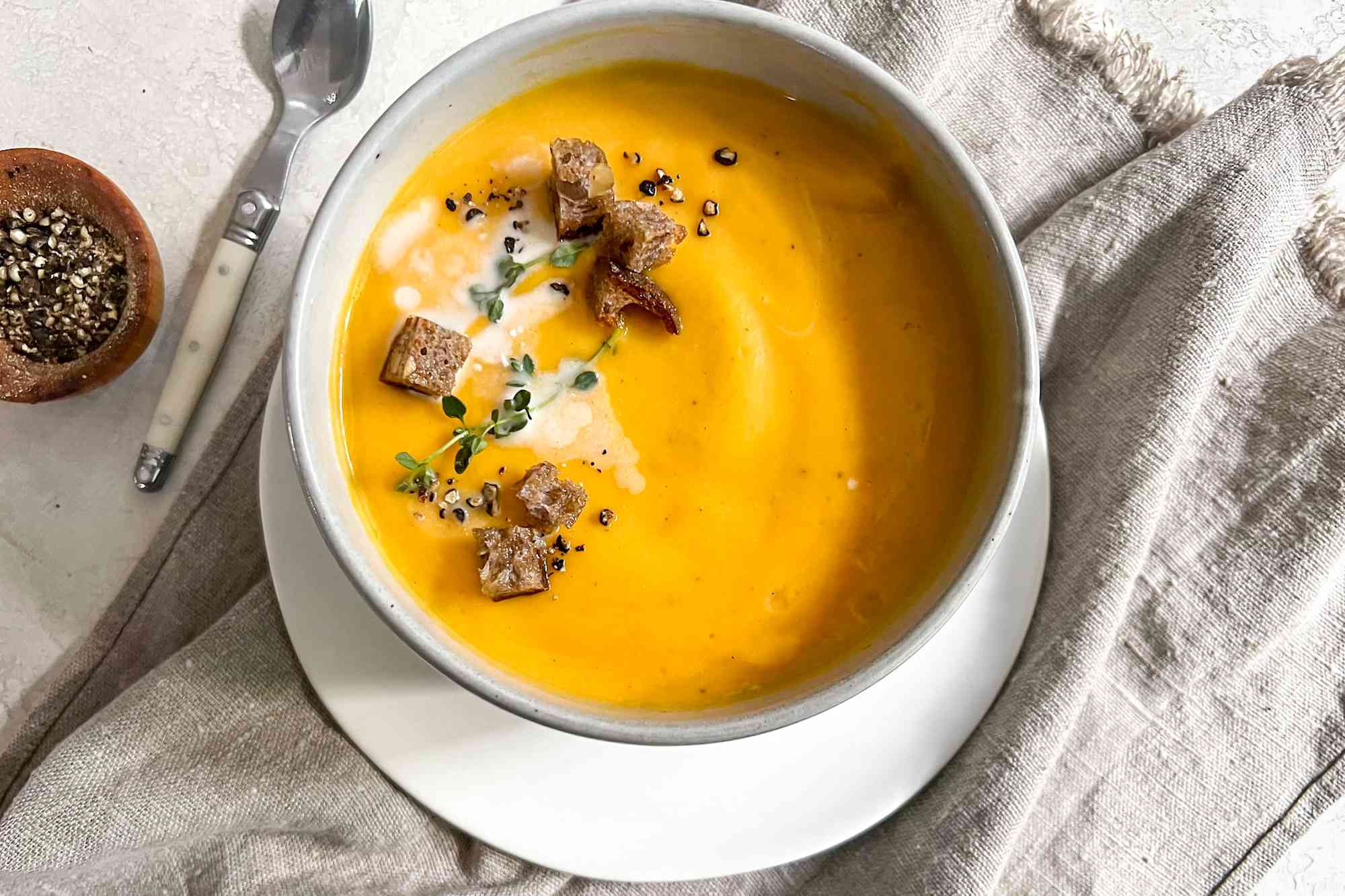 Bowl of Vegan Butternut Squash Soup Topped with Thyme, Coconut Milk, Croutons, and Cracked Pepper, Sitting on a Kitchen Linen and Surrounded by a Bowl of Cracked Pepper and a Spoon 