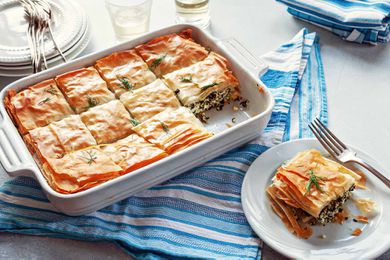 Slice of Greek Spinach Pie on a white plate, with a fork, a glass of white wine, and the casserole dish in the background,