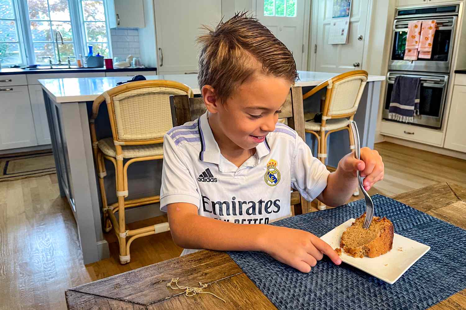A young boy in a Real Madrid jersey eating cake at a kitchen table with a modernstyle kitchen in the background