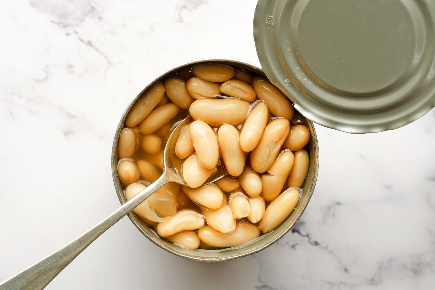 An opened can of white beans with a spoon inside placed on a marble countertop