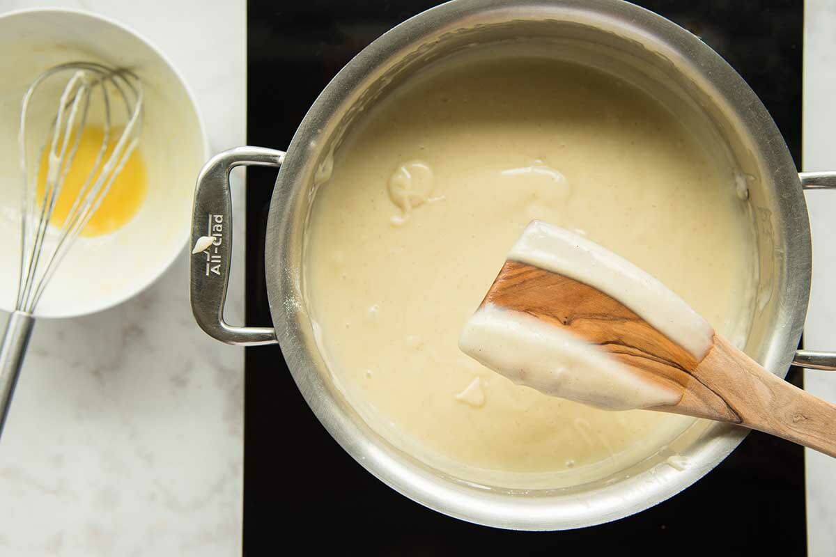 A saucepan of thick custard is on the stove and a wooden spoon is coated in the custard.