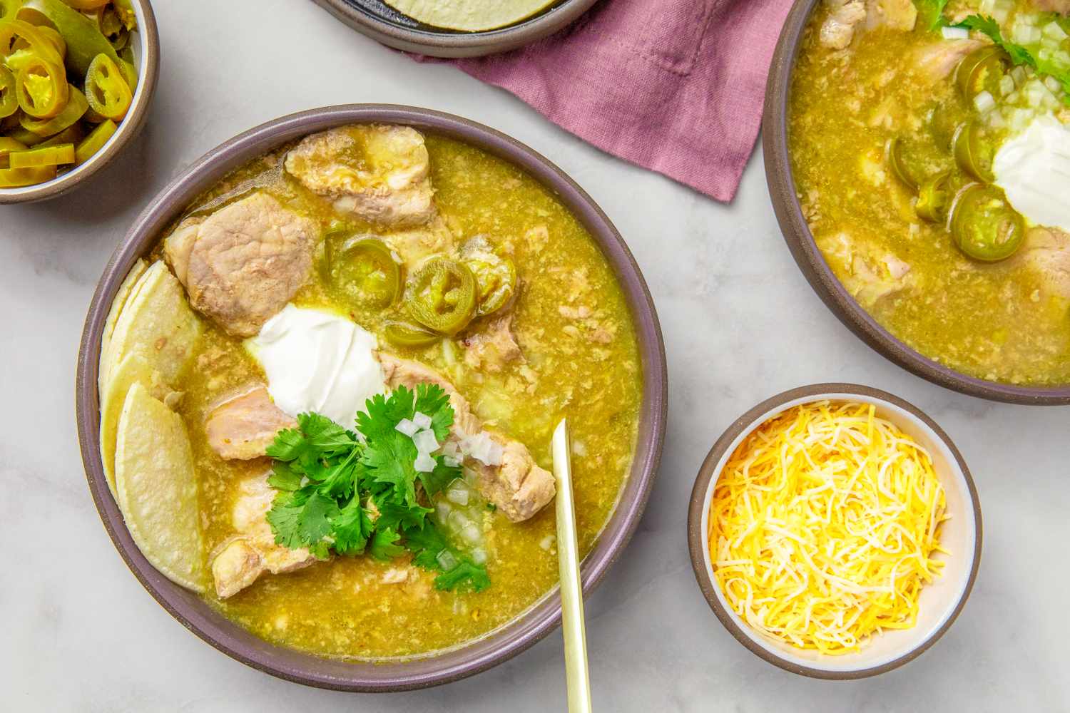 A bowl of pork chile verde garnished with sour cream cilantro and jalapenos accompanied by lime wedges and shredded cheese in bowls