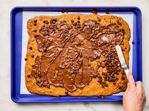 baked flattened cookie on making tray, chocolate chips scattered on top, hand spreading melting chocolate with a small spatula